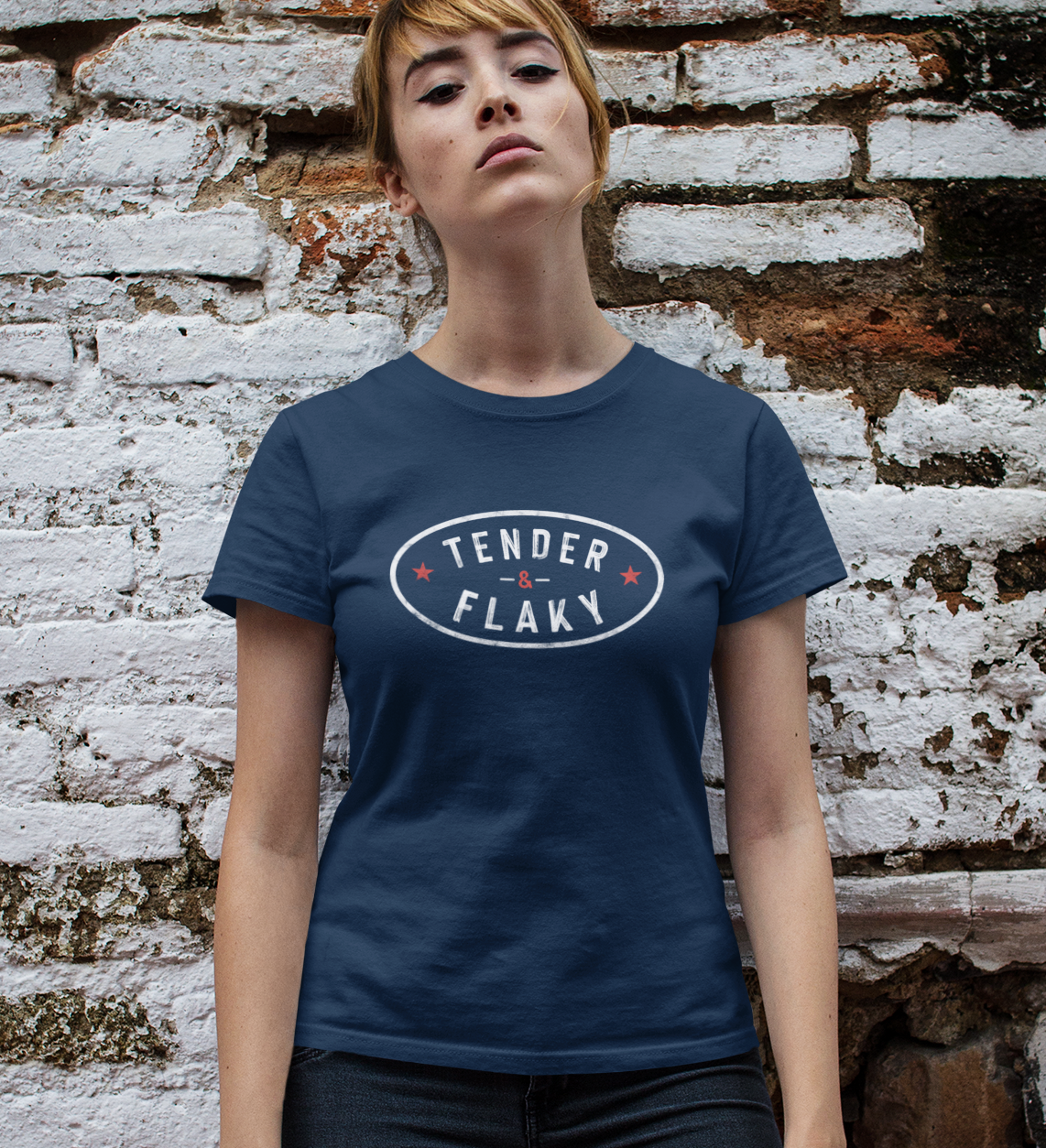 Person wearing a navy blue t-shirt with 'TENDER FLAKY' text against a textured brick wall.